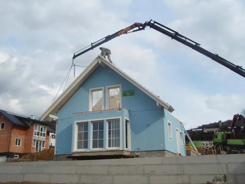 Ein Kran hebt ein Dachteil auf ein hellblaues Haus, auf dem First steht ein Dachdecker