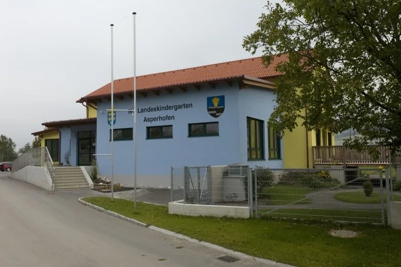 Landeskindergarten Asperhofen mit rotem Ziegeldach, davor Fahnenmasten und die Zufahrt